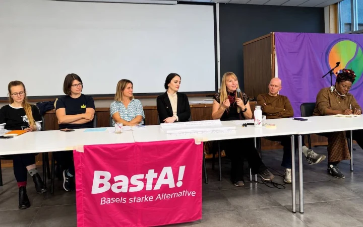 Basel’de BastA! etkinliğinde konuşmacılar ve katılımcılar uzun bir masada oturuyor; masanın önünde BastA! yazılı pembe pankart bulunuyor.