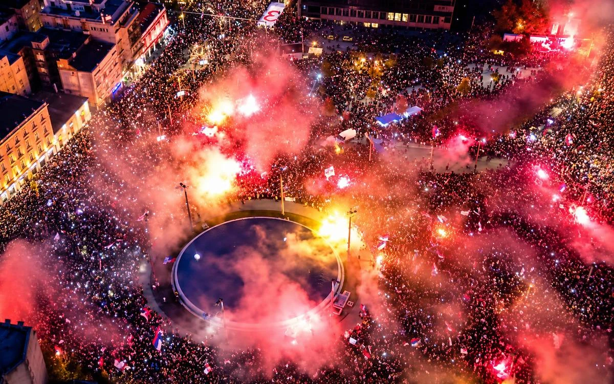 Belgrad’da gece düzenlenen mitingde kalabalık Slavija Meydanı çevresini doldururken kırmızı işaret fişeklerinden dumanlar yükseliyor.