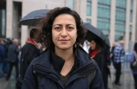 Gazeteci Derya Okatan, arka planda şemsiyeler ve bir bina görülürken kameraya bakıyor.