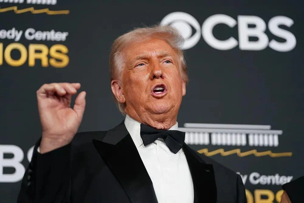 Donald Trump, siyah smokinle bir etkinlikte konuşurken elini kaldırıyor.