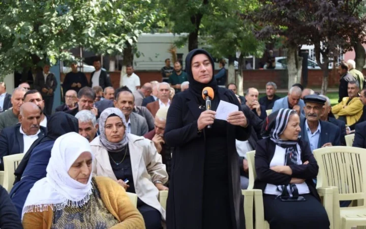 Emine Teğin, açık havada düzenlenen etkinlikte mikrofonla konuşma yapıyor; izleyiciler sandalyelerde oturuyor.