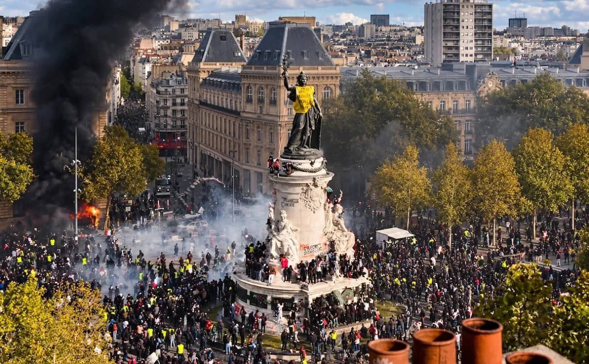 Paris’te bir meydanda toplanan göstericiler, heykel çevresinde ve çevredeki sokaklarda görülüyor; sol tarafta yoğun siyah duman yükseliyor.