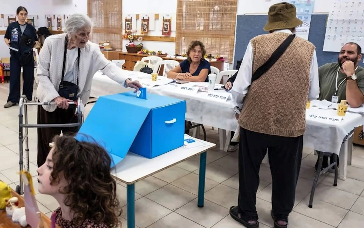 İsrail’de bir seçim merkezinde seçmen oyunu sandığa atıyor; görevli ve diğer seçmenler masaların çevresinde bulunuyor.