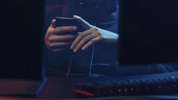 Karanlık ortamda akıllı telefon kullanan bir kişinin elleri ve ön planda bilgisayar klavyesi