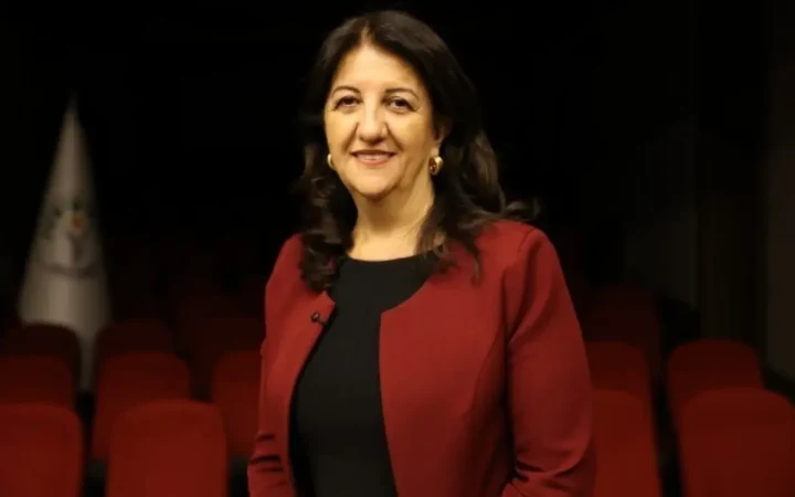 Pervin Buldan kırmızı ceketle, kırmızı koltukların bulunduğu bir salonda ayakta duruyor.