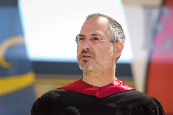 Steve Jobs, gözlüklü ve kırmızı-siyah akademik cübbe içinde bir etkinlikte konuşurken
