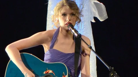 Taylor Swift, gitarıyla sahnede mikrofonla şarkı söylüyor.