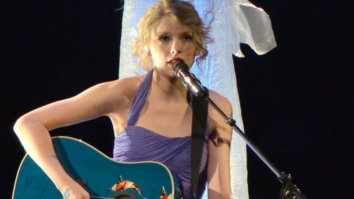 Taylor Swift, gitarıyla sahnede mikrofonla şarkı söylüyor.