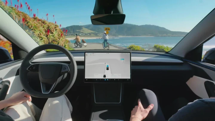 Tesla otomobilinin içinden görülen kokpit, direksiyon ve merkezi ekran; ön camın dışında bisikletliler bulunuyor.