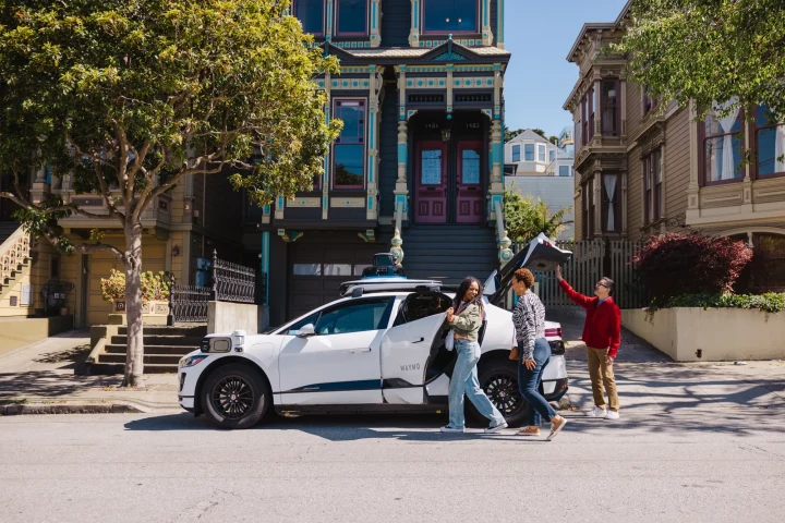 Beyaz bir robotaksinin yanında duran üç kişi, San Francisco’daki bir sokakta görülüyor.