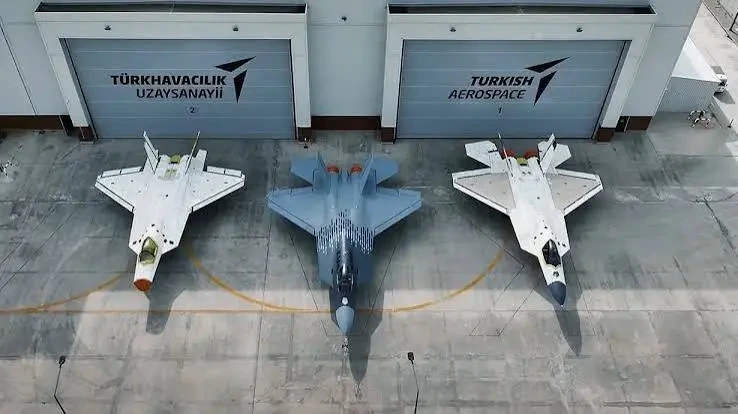TUSAŞ hangarlarının önünde üç hava aracı; ortada koyu renkli, iki yanında beyaz uçaklar bulunuyor.